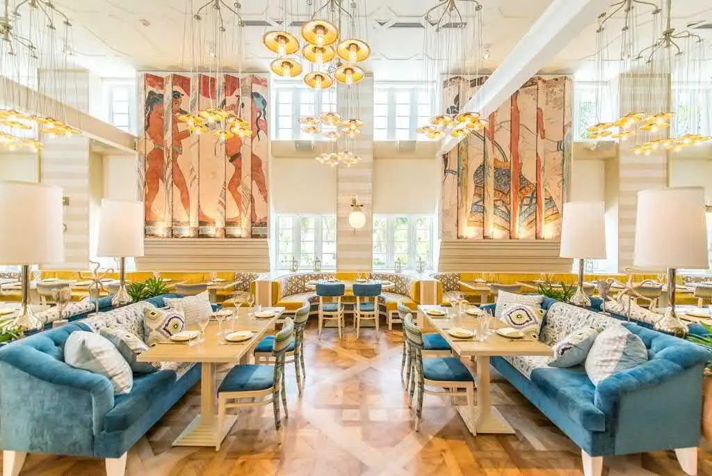Byblos Miami – Elegant Mediterranean Dining Room A bright Mediterranean-style dining room at Byblos Miami, featuring blue and white patterned curtains, coral and beige seating, and elegant modern chandeliers. Tables are set with fine glassware, plates, and folded napkins.
