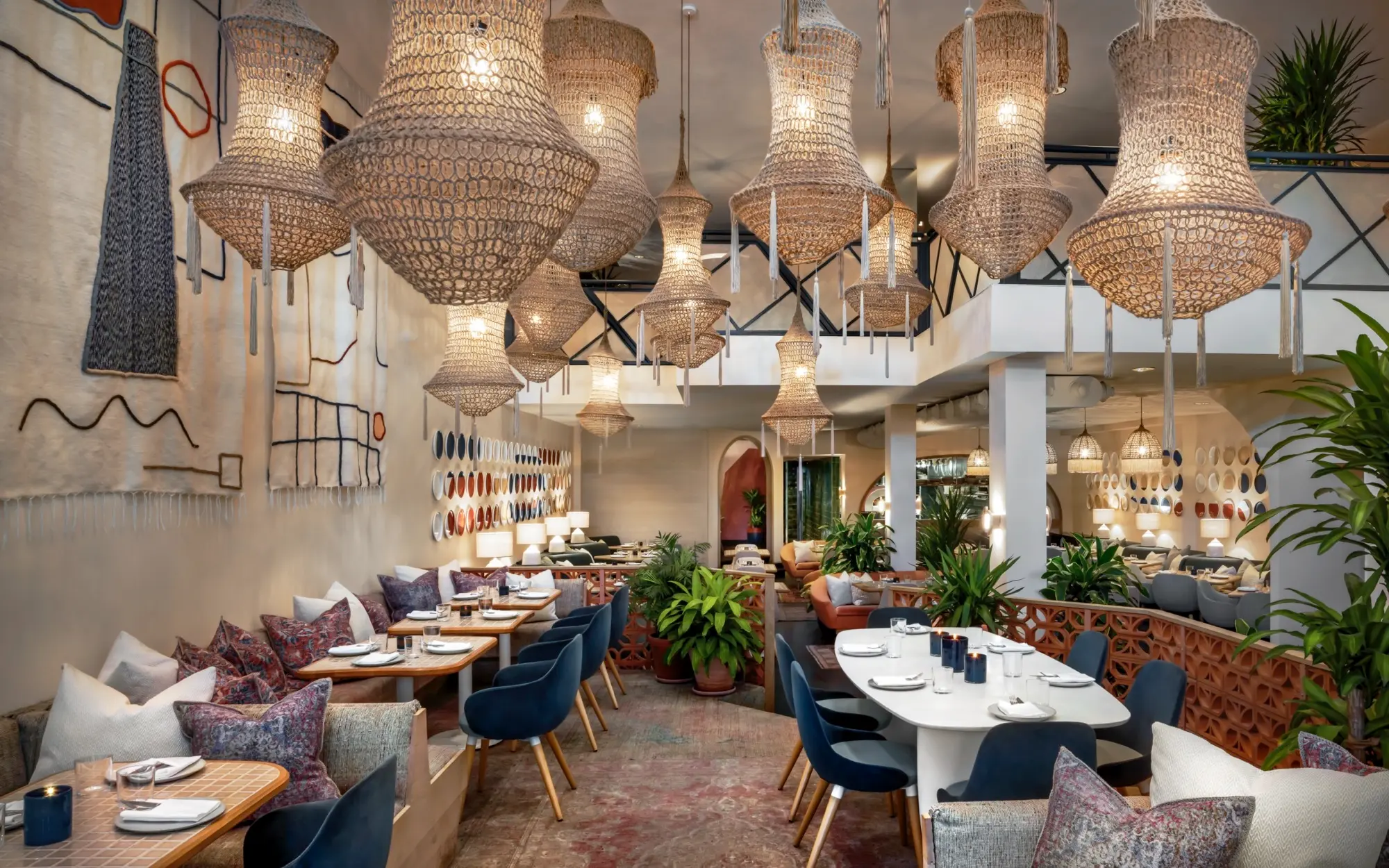 Byblos Uptown – Artisanal Mediterranean Dining Byblos Uptown’s dining area featuring handcrafted woven chandeliers, earthy-toned seating, arched architectural elements, and lush greenery. The walls display artistic patterns with Mediterranean textures.