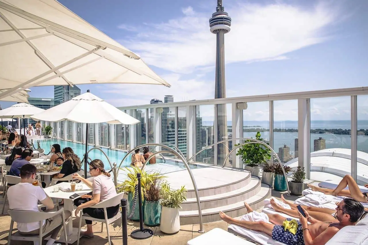 Kōst Rooftop Patio with CN Tower View A luxurious rooftop patio at Kōst, overlooking the Toronto skyline with the CN Tower in the background. Guests are dining and relaxing by the infinity pool under large white umbrellas.